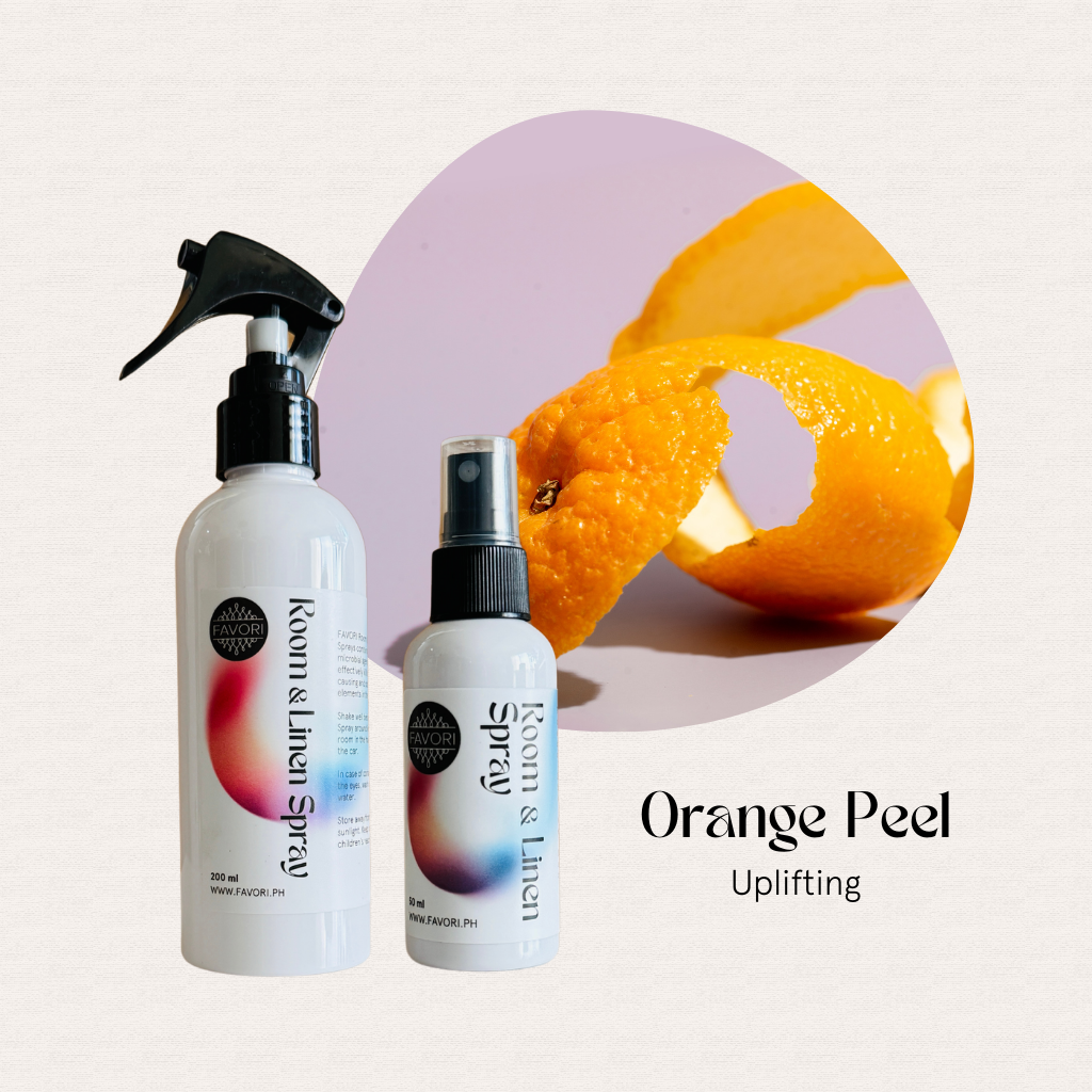 Two FAVORI Scents Regular Room & Linen Air Sprays sit beside a curled orange peel on a light background.