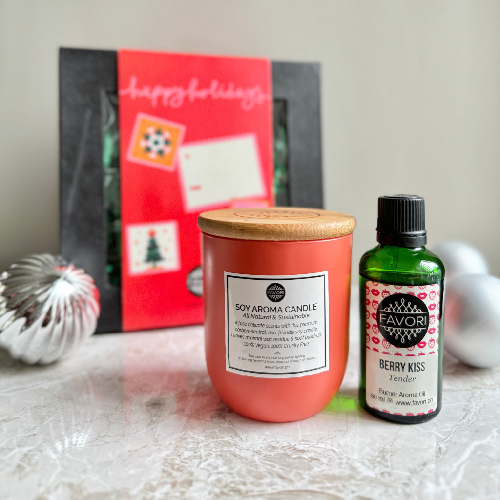 A soy aroma candle from FAVORI Scents, in a peach-colored jar, sits on a marble surface next to an Aroma Oil from the Burner collection labeled "FAVORI Berry Kiss." Behind them, there is a black gift package adorned with a red holiday greeting card. Two silver textured ornaments are partially visible to the side.