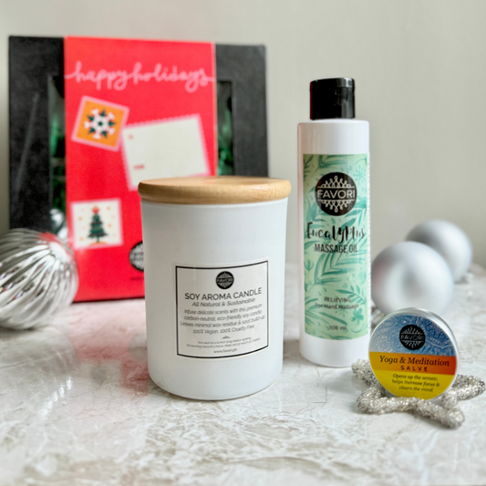 Displayed on a marble surface is the FAVORI Scents holiday-themed relaxation gift package. It features a white 160g soy aroma candle with a wooden lid, a 100ml bottle of eucalyptus massage oil, and a 15g wellness salve. In the background, there's a red gift box adorned with festive decorations, complemented by a silver ornament.