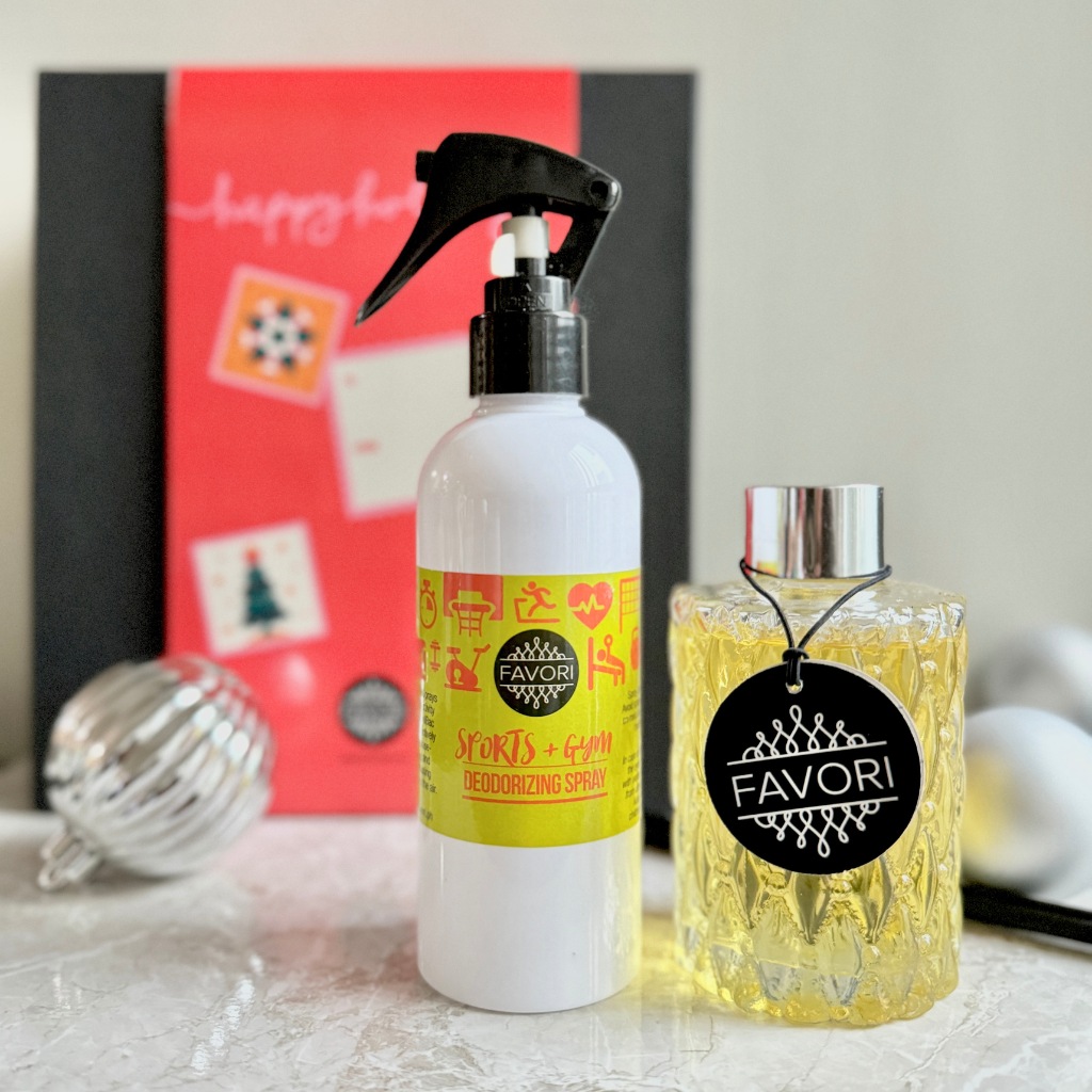 A white spray bottle from the "Air Spray (200ml) + Premium Reed Diffuser (150ml)" set featuring yellow graphics sits beside a decorative reed diffuser labeled with the "FAVORI Scents" brand. In the background, part of a red-and-black box is visible, with a metallic ornament resting on the marble surface.