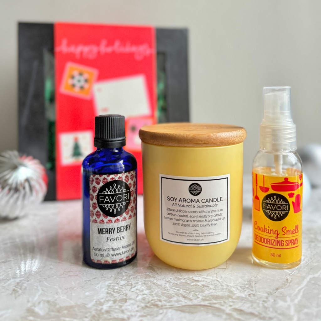 On a table is a holiday-themed gift set from FAVORI Scents, featuring an Aerator/Diffuser (AD) Aroma Oil in blue packaging, a yellow 160g soy aroma candle, and an air spray labeled "Cooking Smell Deodorizing." In the background, you can see a red holiday card with blurred text and a partially hidden silver ornament.