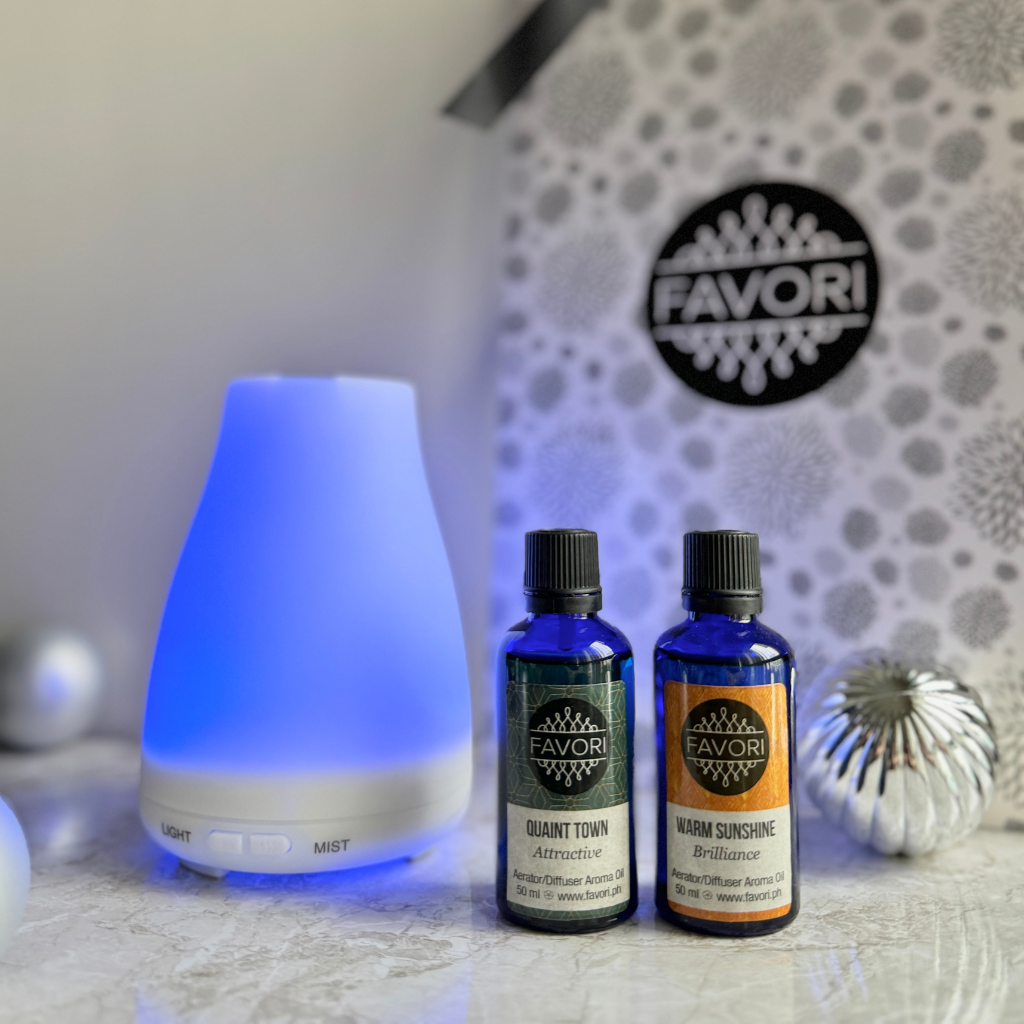 A Vim Diffuser from FAVORI Scents gently casts a soft blue glow, accompanied by two bottles of 50ml aroma oils named "Quaint Town" and "Warm Sunshine," which sit gracefully on a marble surface. In the background, a floral-patterned decorative gift bag and an ornamental silver sphere complete this serene gift package.