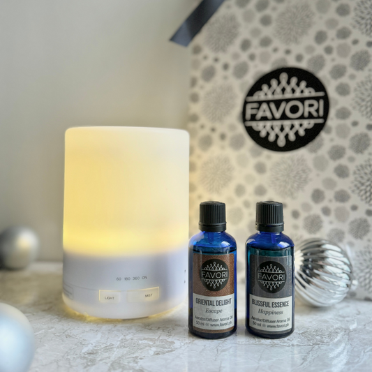 A Lind diffuser emits a gentle white glow next to two blue 50ml bottles of essential oils labeled "Oriental Delight" and "Blissful Essence." The elegant FAVORI Scents gift package is showcased in the background, with decorative silver spheres adding to the charm of these aromatic oils, all beautifully displayed on a marble surface.