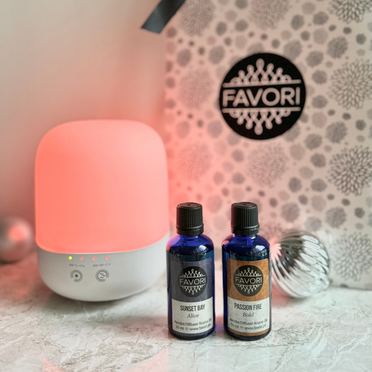 A soft pink Capsule Diffuser from FAVORI Scents casts a warm glow on a marble surface. In front, two blue FAVORI aroma oil bottles labeled "Sunset Bay" and "Passion Fire" stand with a decorative pattern in the background, accompanied by a reflective silver ornament. A GRANDE gift package bag peeks into view.