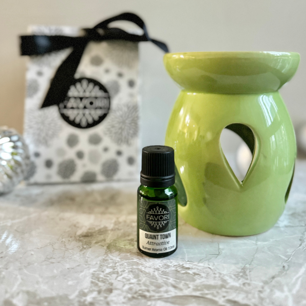 A Raindrop Ceramic Burner Device from FAVORI Scents, with a green finish and heart-shaped cutouts, is placed on a marble surface. In front of it sits a small green bottle labeled "FAVORI Scents - Raindrop Ceramic Burner Device+10ml Burner (BR) Aroma Oil," adding to the scene's aromatic appeal. A decorative bag adorned with a black ribbon and floral pattern enhances the charm of the setting.