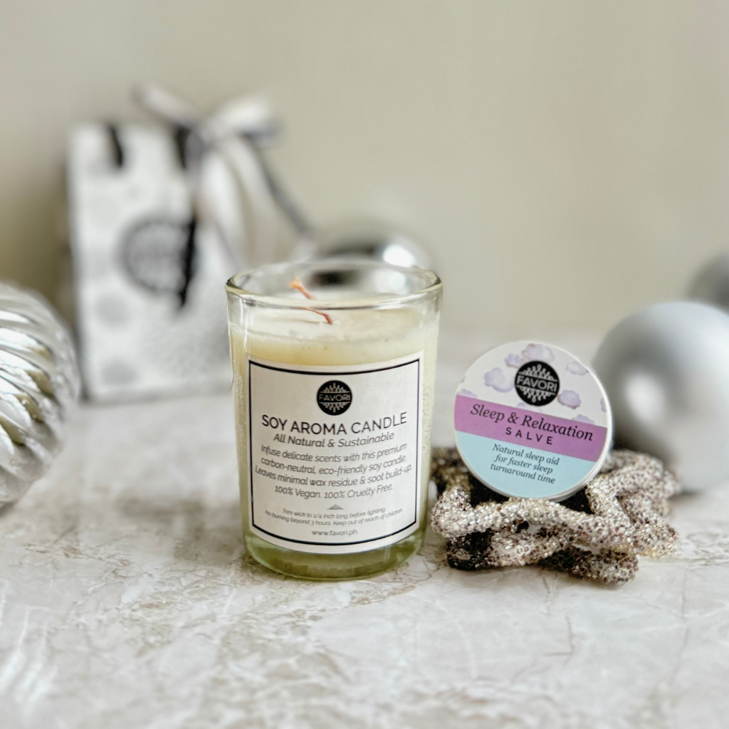 A 60g FAVORI Scents soy aroma candle, adorned with a white label, stands on a textured surface, radiating tranquility. Beside it sits a small 15g wellness salve container labeled "Sleep & Relaxation," infused with lavender and cedarwood fragrance. Blurred in the background is the Relaxation Package, featuring an elegant gift bag and gleaming silver ornaments.